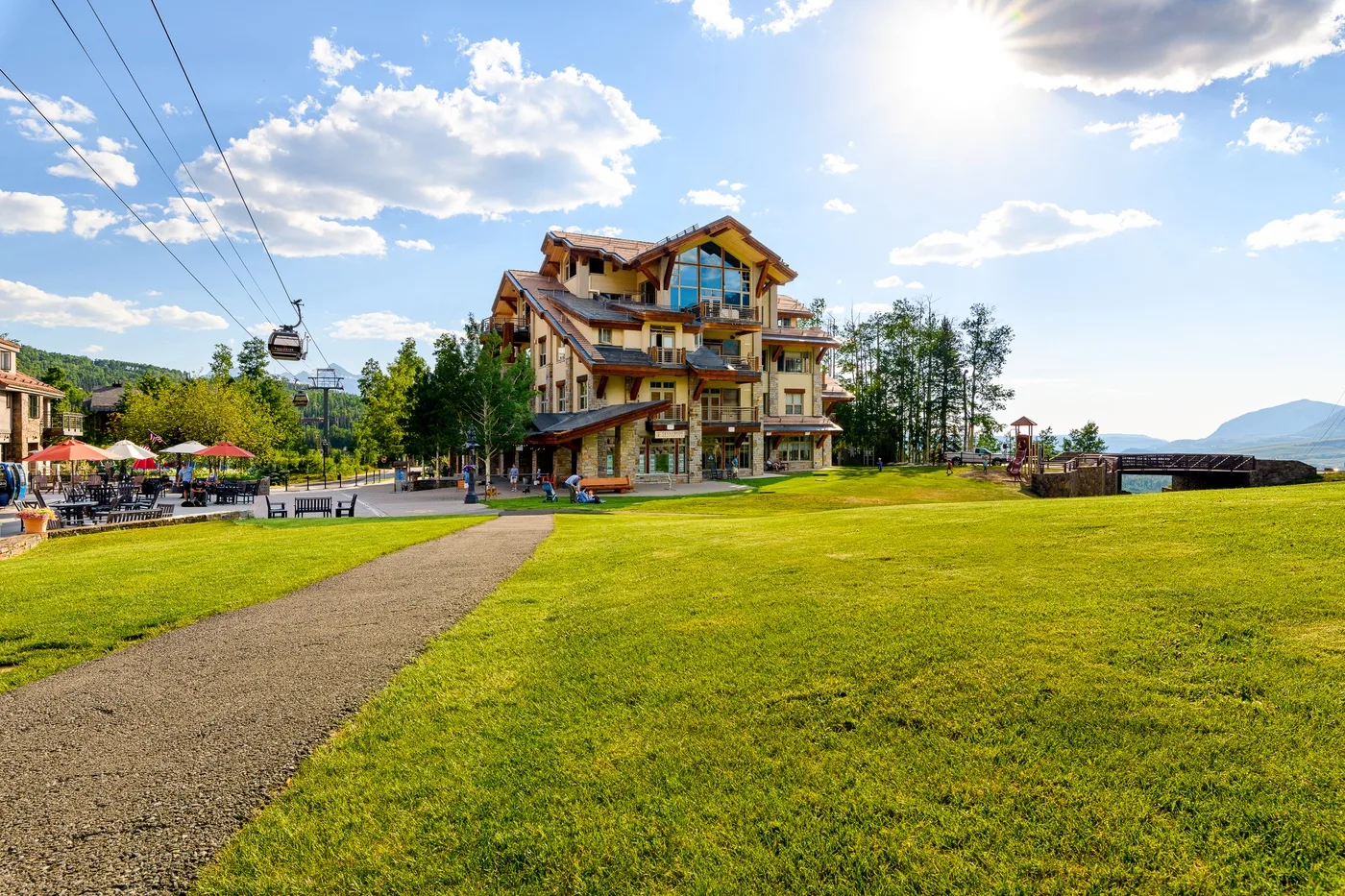A lodge stands beyond a grassy lawn, with a paved path and cable cars nearby.