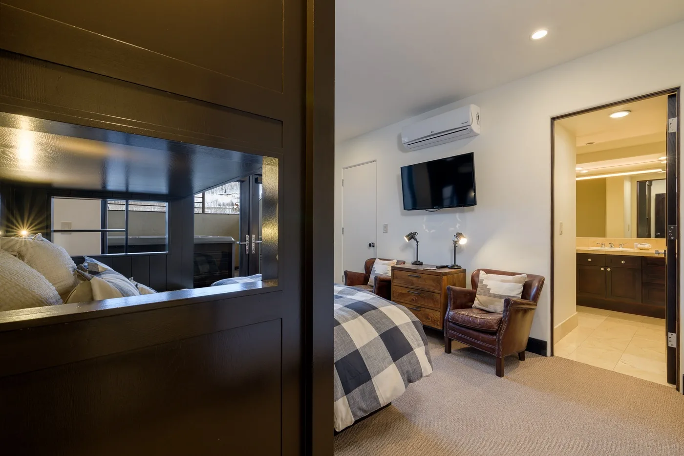 A bedroom with a bed, seating, and a wall-mounted television beside an open bathroom.