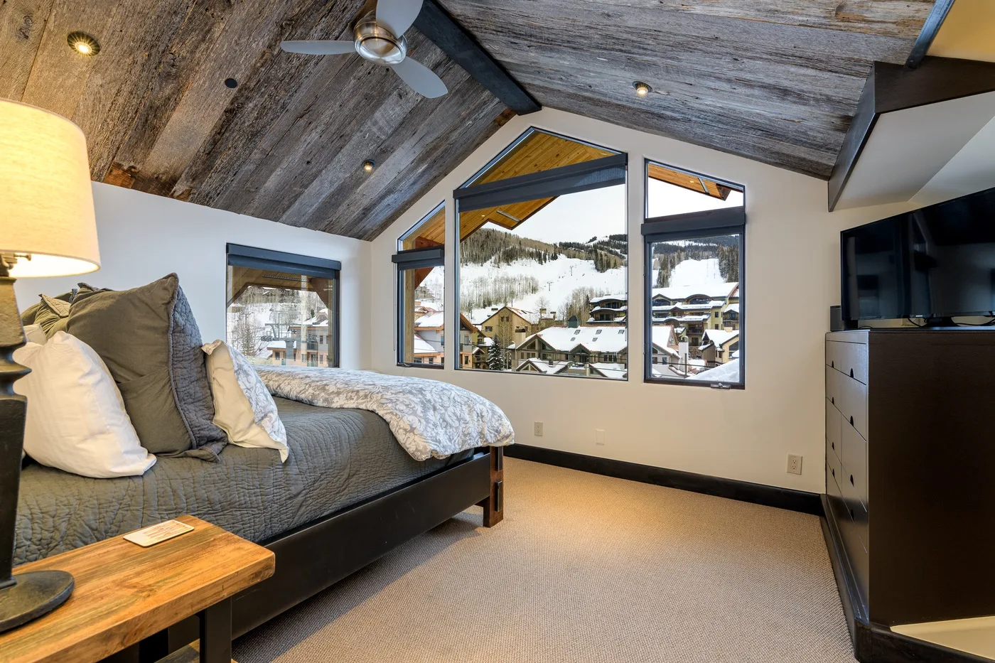 A bedroom with a bed beneath a sloped wood ceiling and windows overlooking a snowy village.