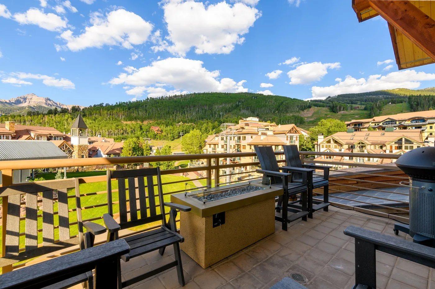 A sunny balcony with outdoor seating overlooks a green hillside and nearby buildings.