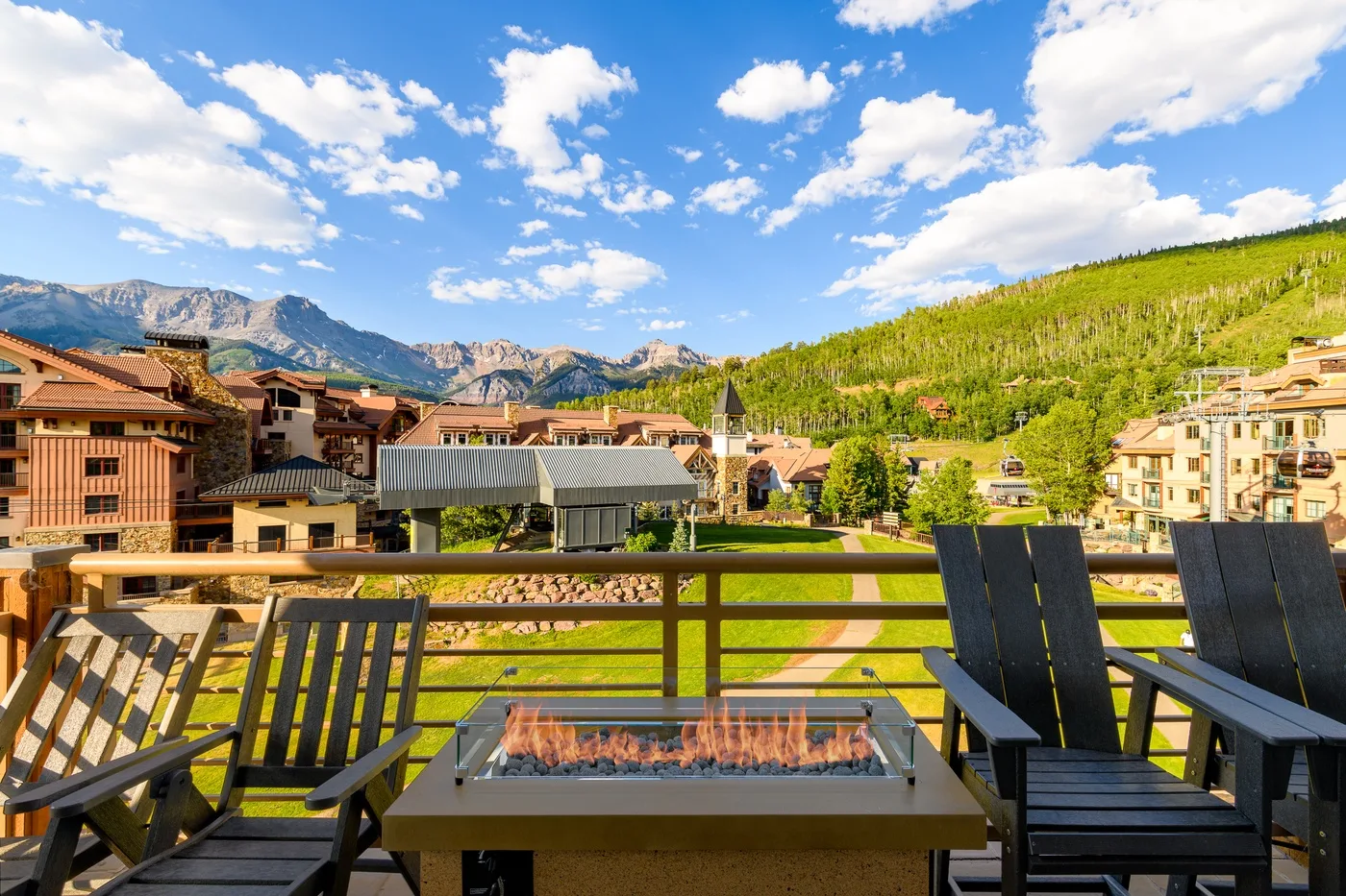Outdoor seating area with a fire table overlooks buildings and green hills.