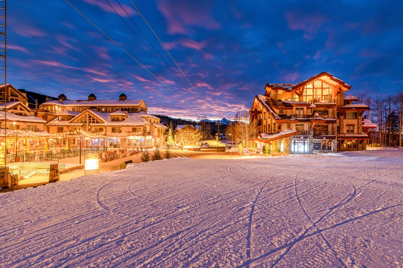 Snow-covered ground stretches between illuminated buildings beneath a colorful evening sky.
