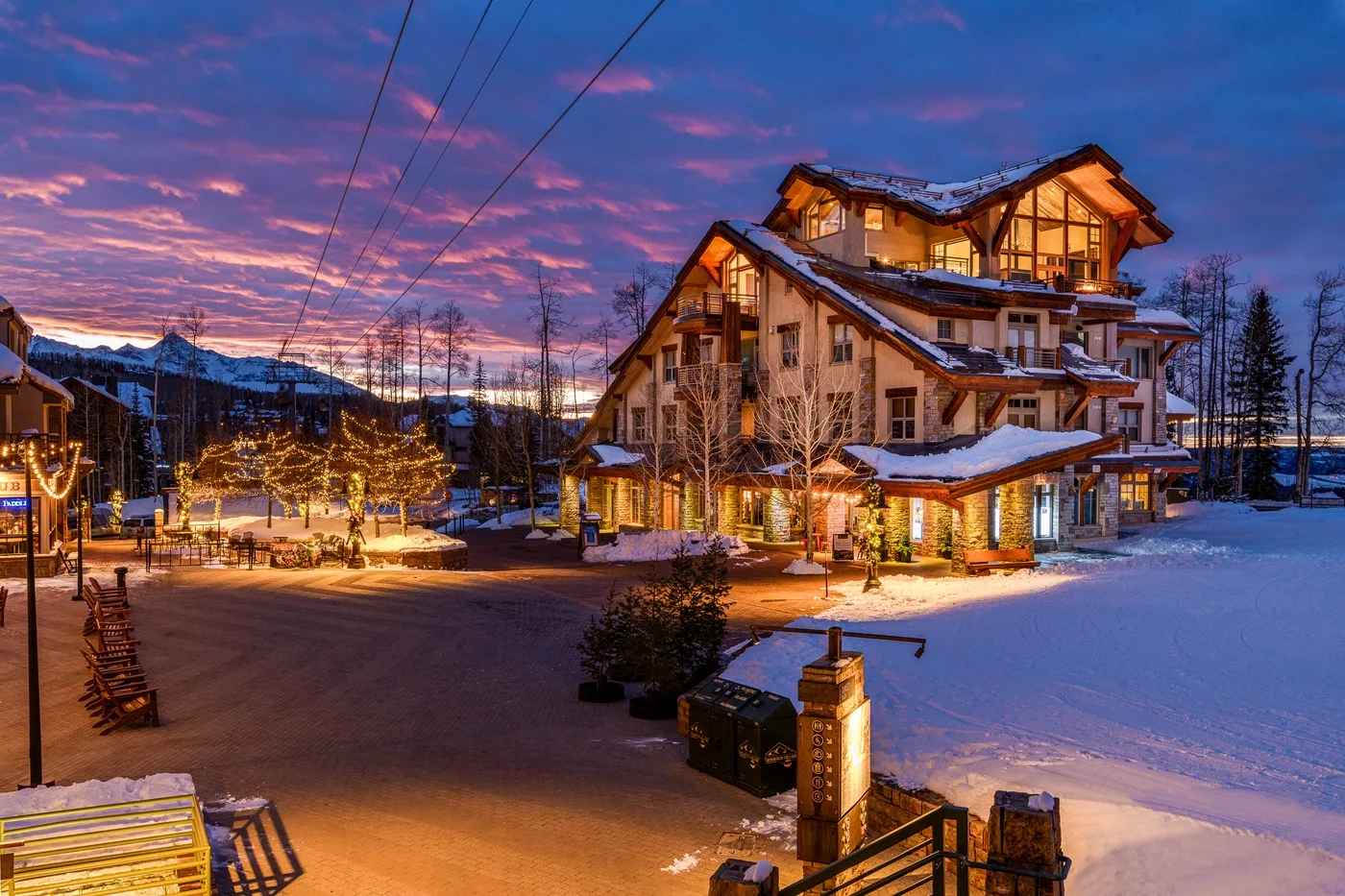 A warmly lit lodge stands beside a snowy outdoor courtyard at dusk.