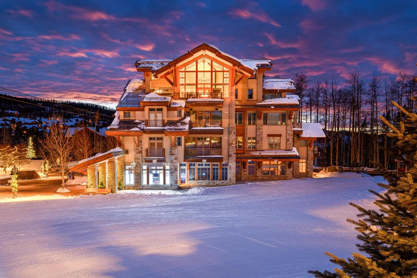 A large illuminated building stands beside a snow-covered open area at dusk.