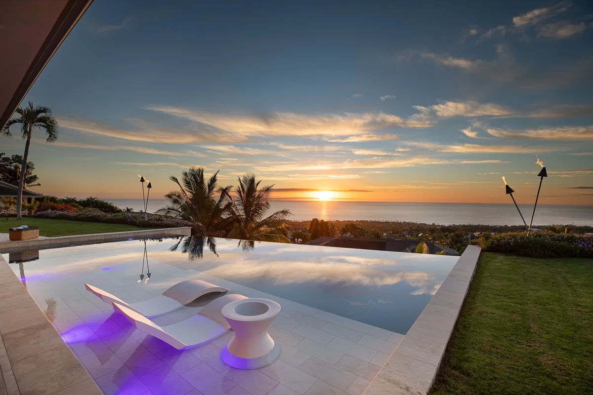 Una piscina al aire libre refleja el atardecer junto a un césped y palmeras.