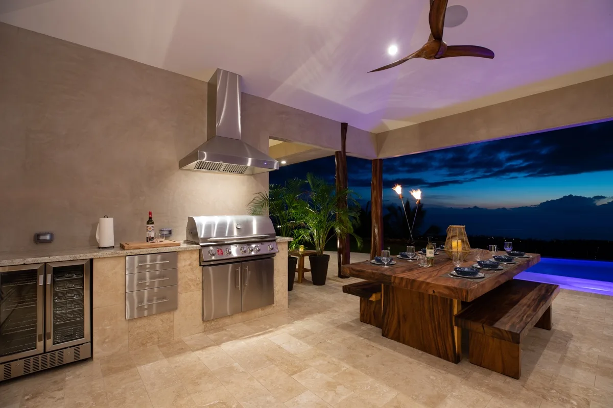 Una zona de comedor al aire libre con una mesa de madera junto a una parrilla, frente al cielo del atardecer.