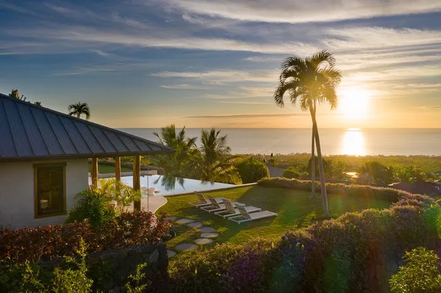 Un jardín costero con palmeras, una piscina y el sol poniéndose sobre el agua.