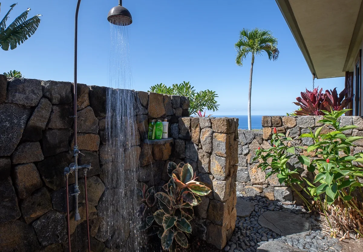 Una ducha exterior está junto a un muro de piedra, con plantas tropicales y vistas al mar.