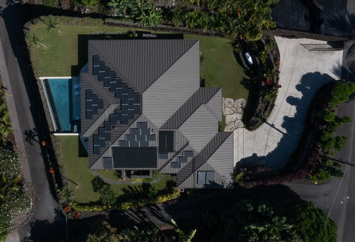 Una vista aérea muestra una casa grande con paneles oscuros en el techo, una piscina y jardines.