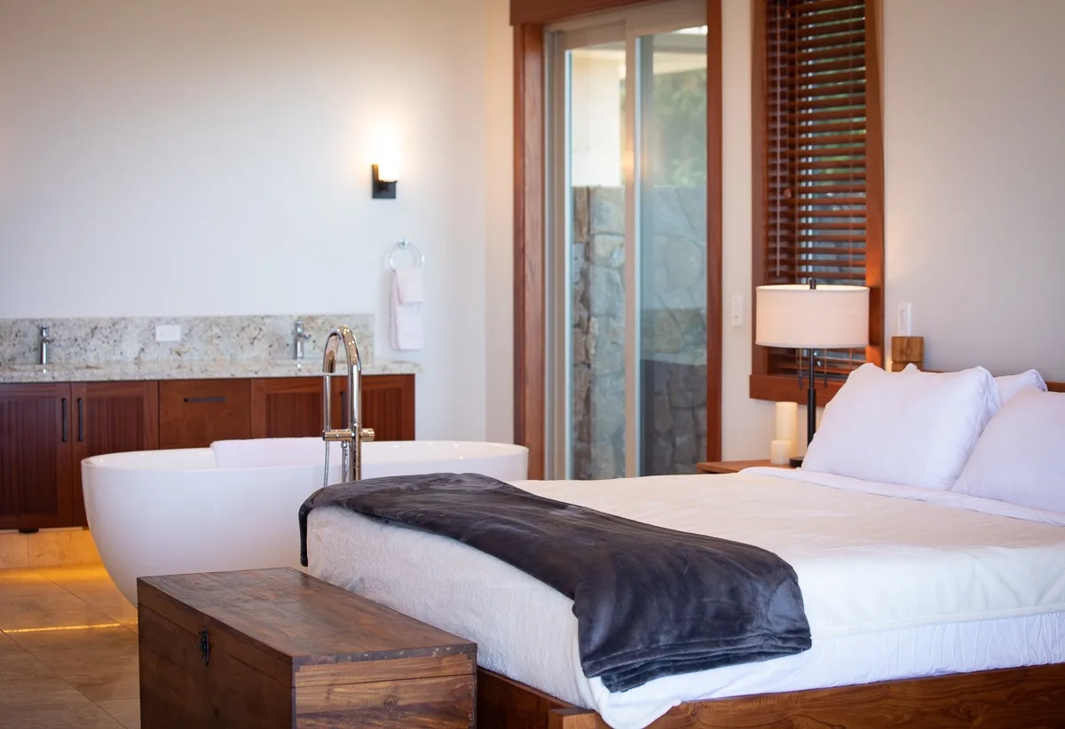 Un dormitorio con una cama blanca, una lámpara de noche y una bañera exenta.