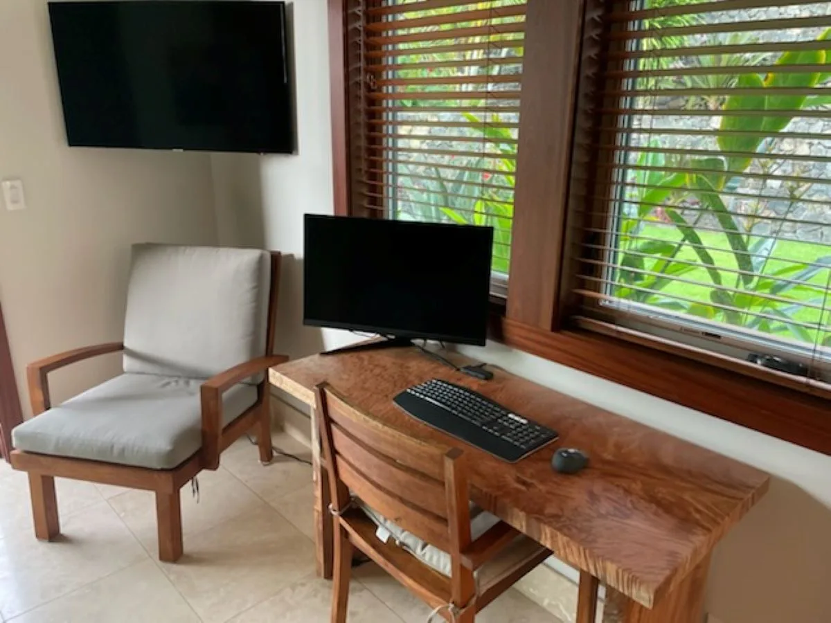 Un escritorio de madera con monitor y teclado está junto a una silla y ventanas con persianas.