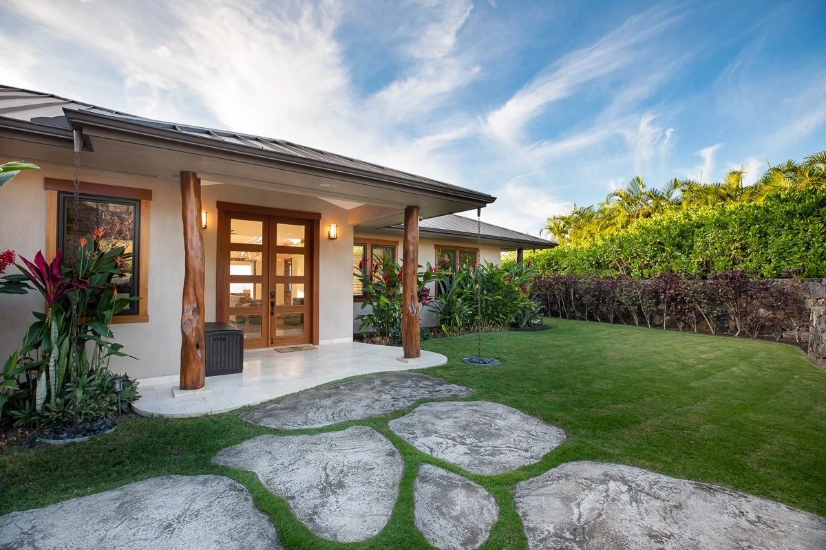 La entrada de una casa da a un césped verde con un camino de piedra y plantas tropicales.