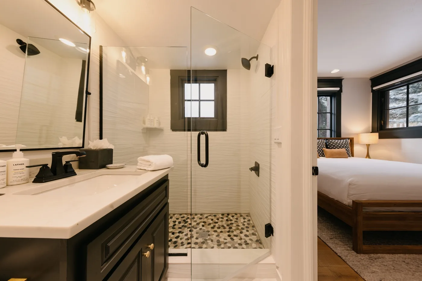 A bright bathroom with a long vanity and a glass shower beside a bedroom.