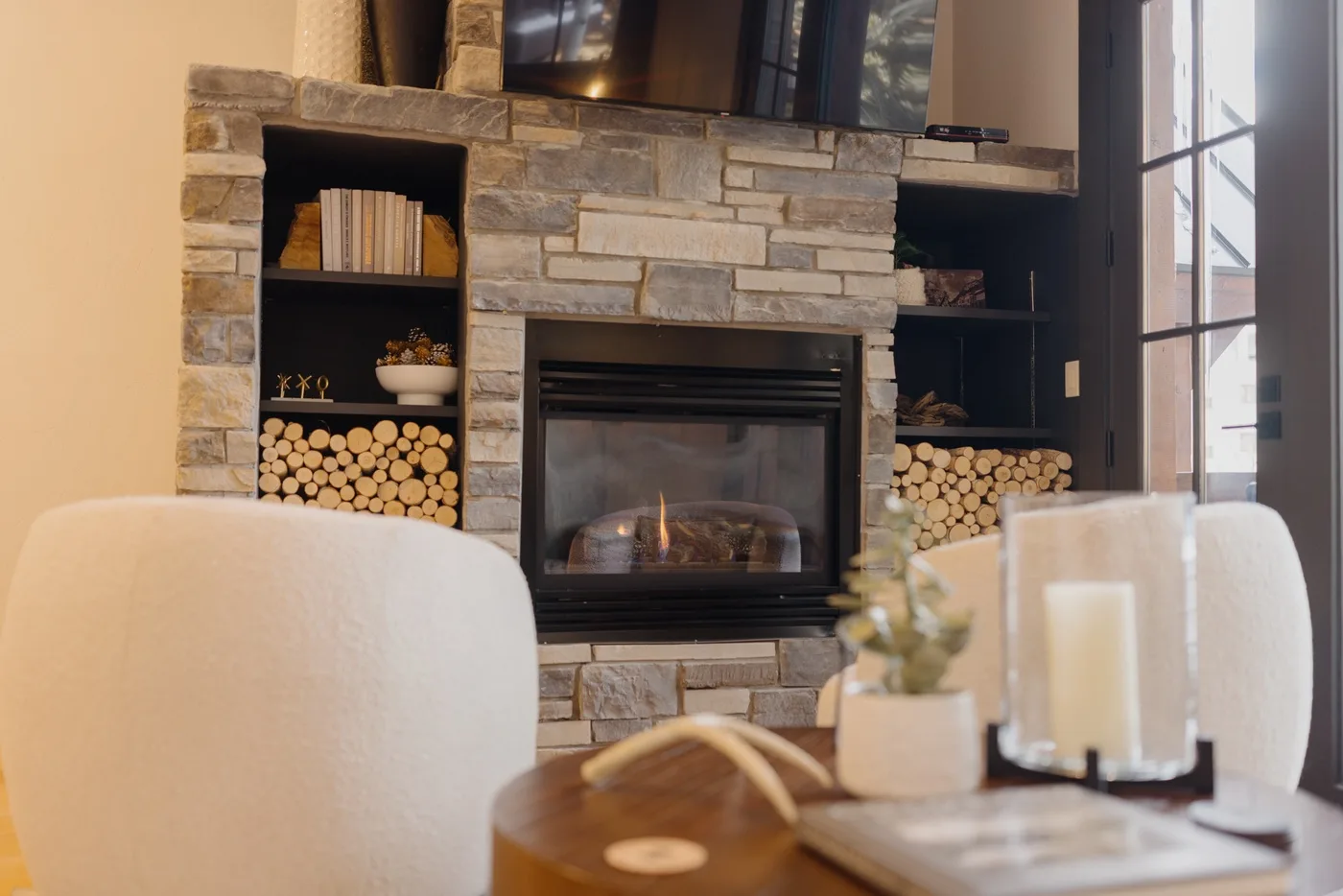 A stone fireplace with stacked logs sits beside a tall window in a living room.