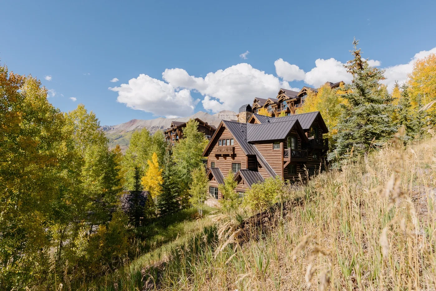A wooden lodge sits among autumn trees on a grassy hillside.