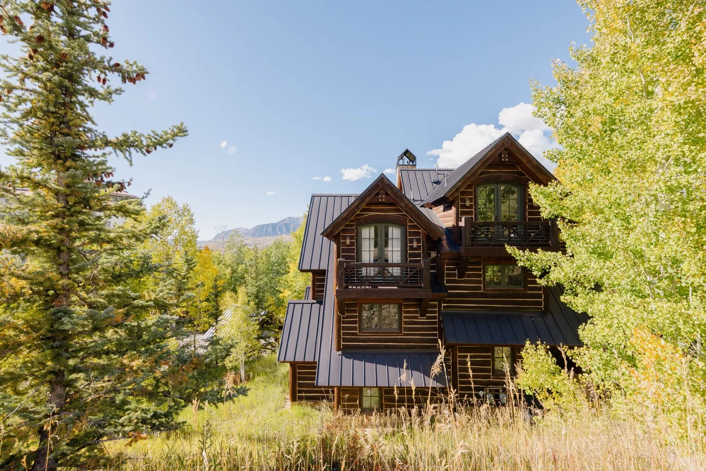 A large wooden lodge stands among trees in a sunny mountain landscape.