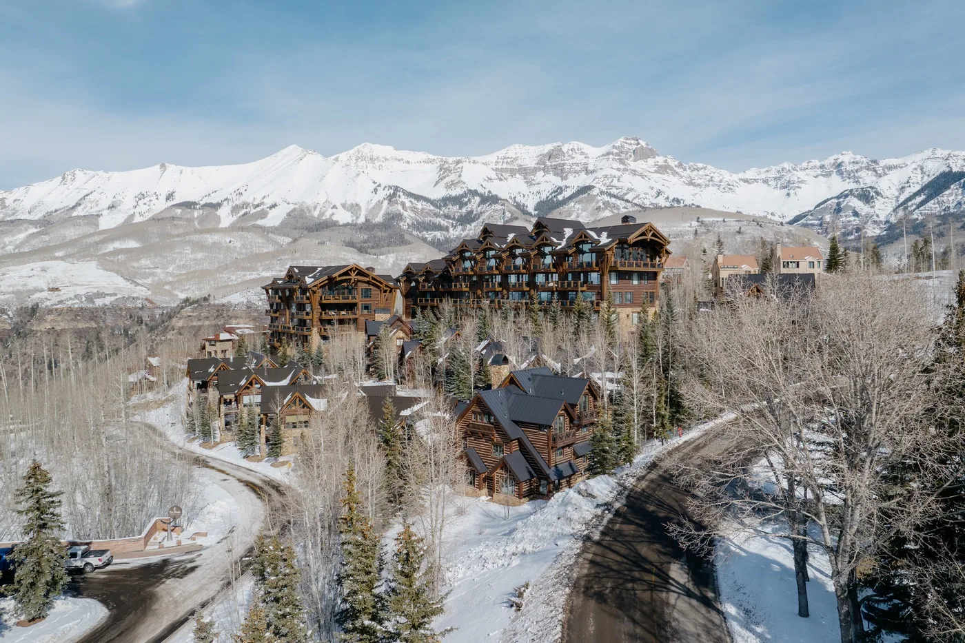A mountain resort complex sits among snowy trees beneath a snow-covered mountain range.