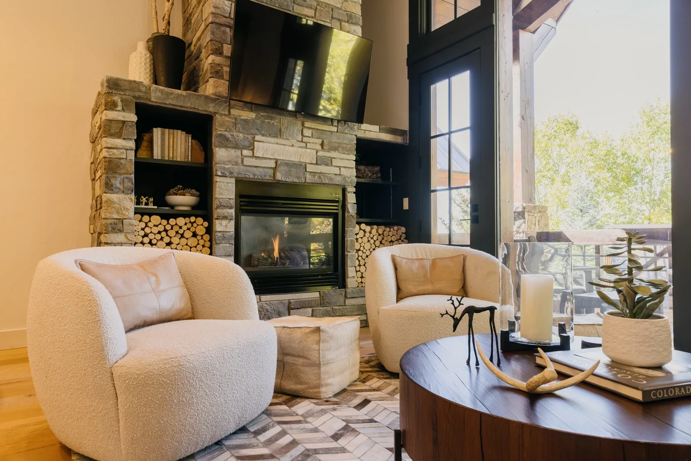 A living room with a stone fireplace, seating, and tall windows overlooking greenery.