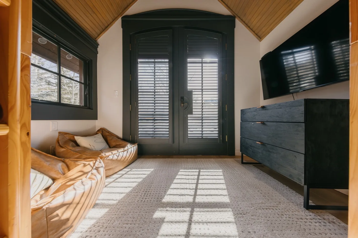 A bright sitting room with cushioned seating, a dark cabinet, and shuttered windows.