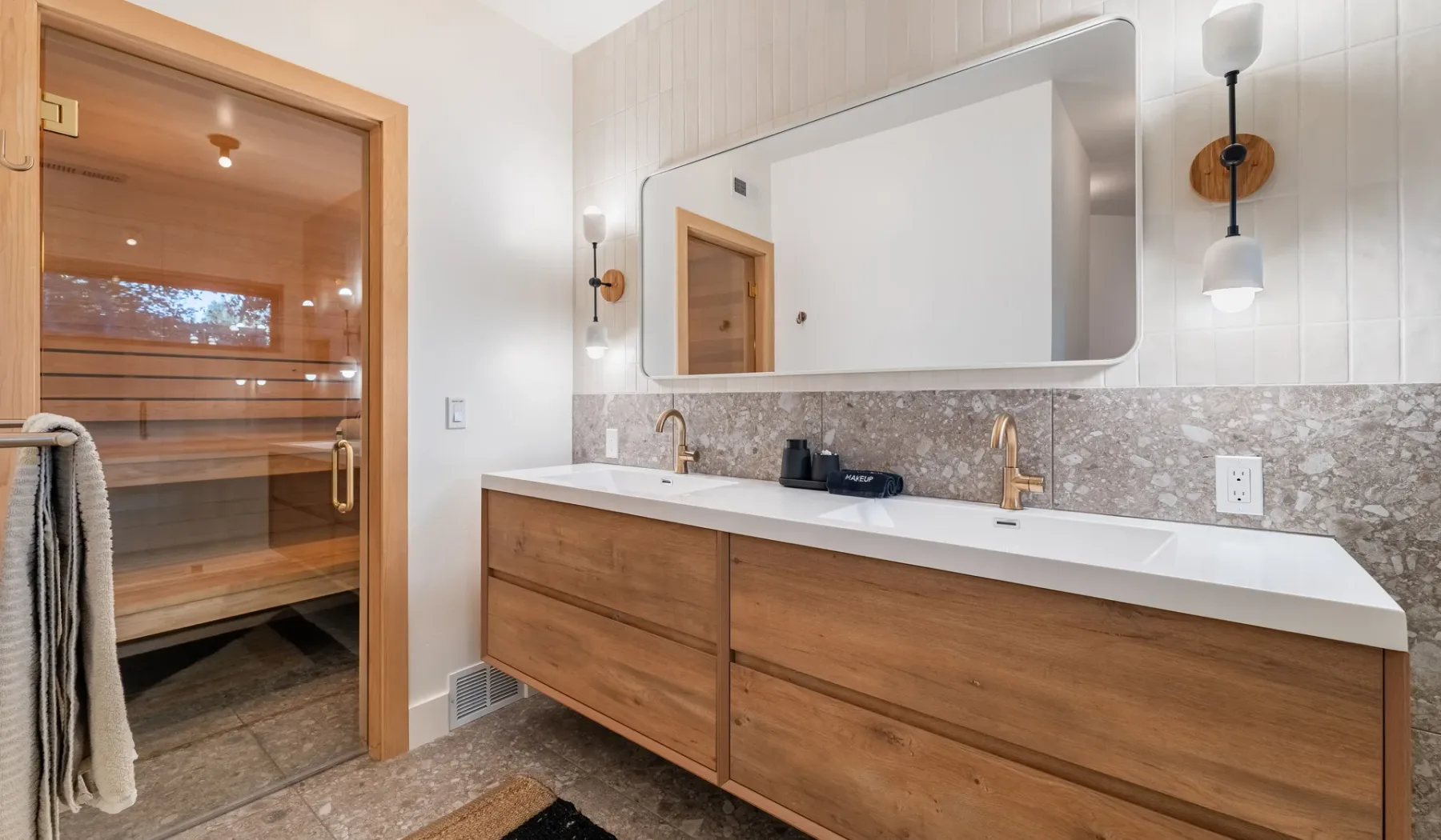 Un mueble de baño con un espejo amplio está junto a una sauna con frente de vidrio.