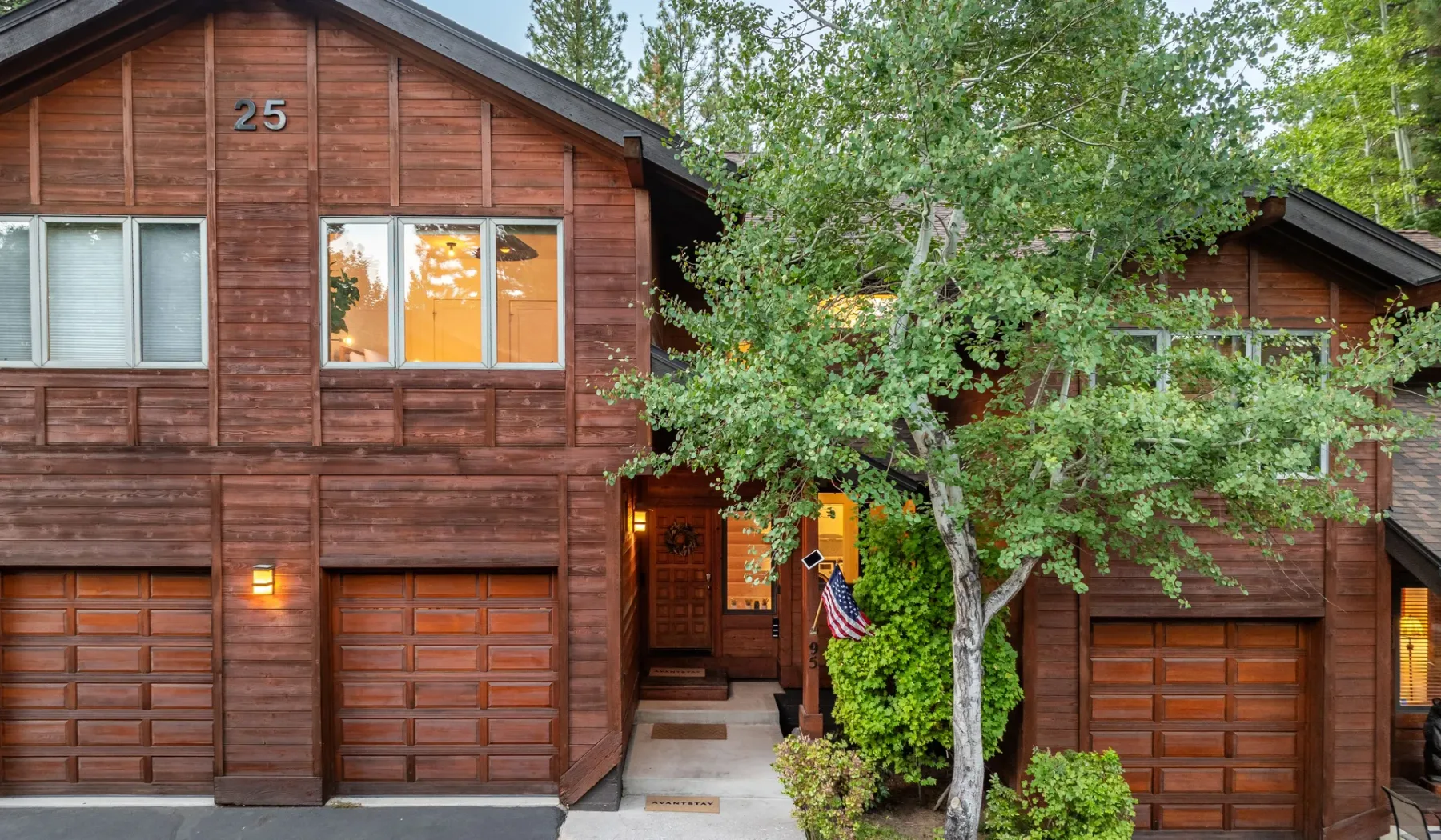 Exterior de una casa de madera con garajes, ventanas y un árbol frondoso delante.