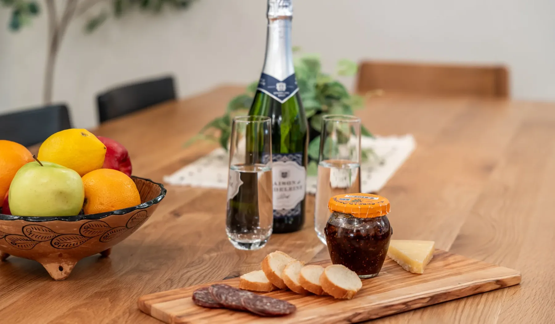 Una mesa de comedor está preparada con fruta, una botella, copas, pan y un frasco.