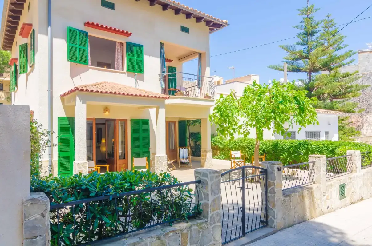 Una casa de dos plantas con contraventanas verdes se alza tras un muro de piedra y un jardín.