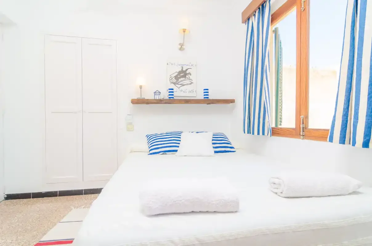 Un dormitorio luminoso con una cama blanca y cojines a rayas azules junto a una ventana.