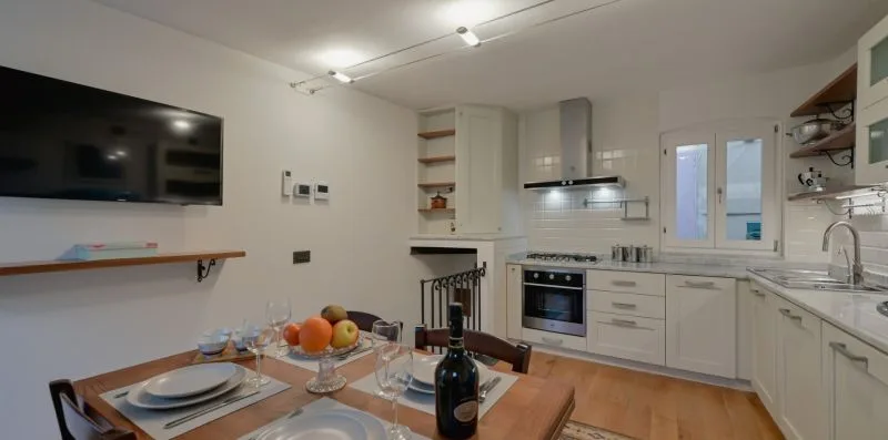 Una cocina luminosa con gabinetes blancos, una mesa de comedor y un televisor en la pared.