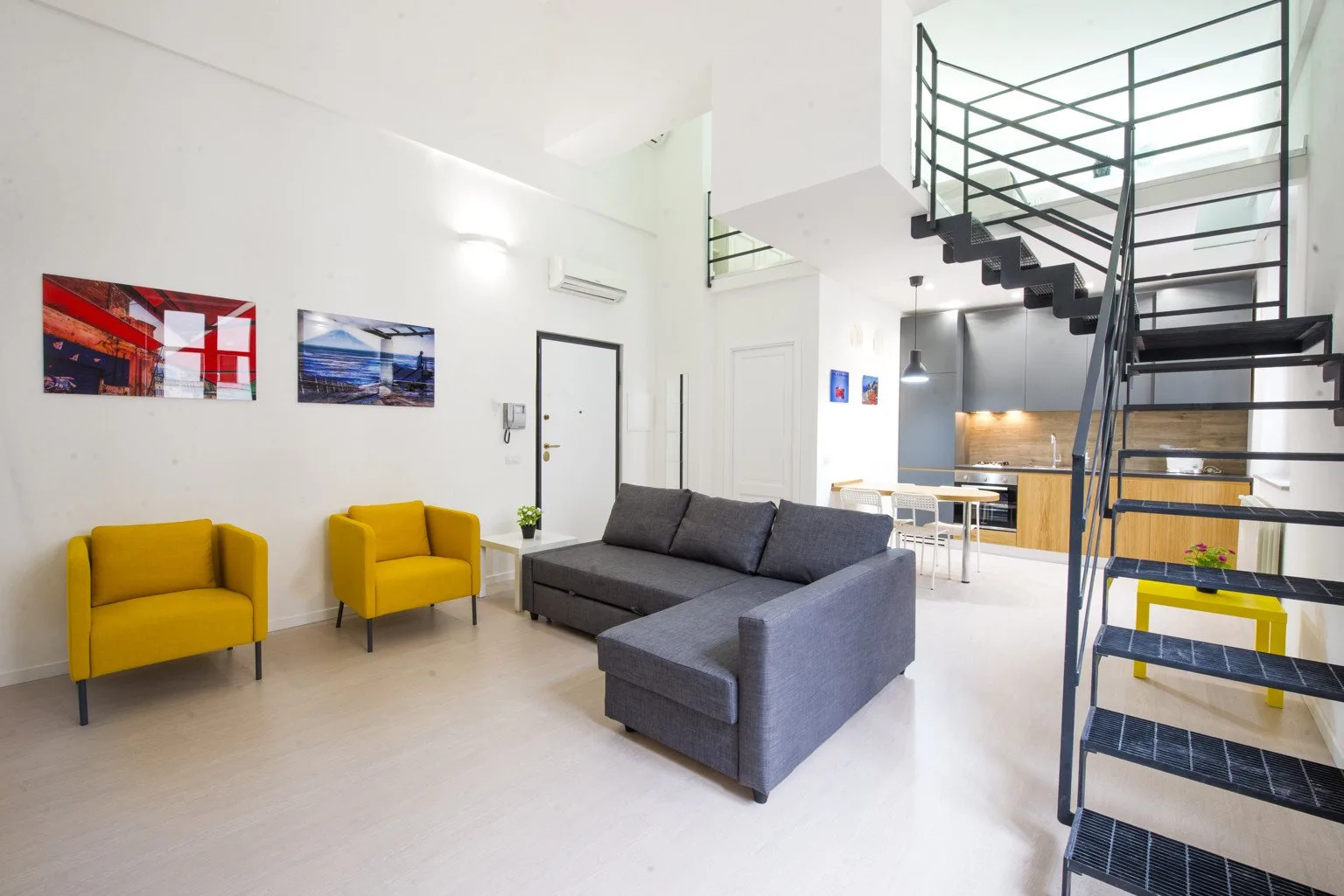 A bright open-plan living area with gray seating, yellow armchairs, and a staircase.