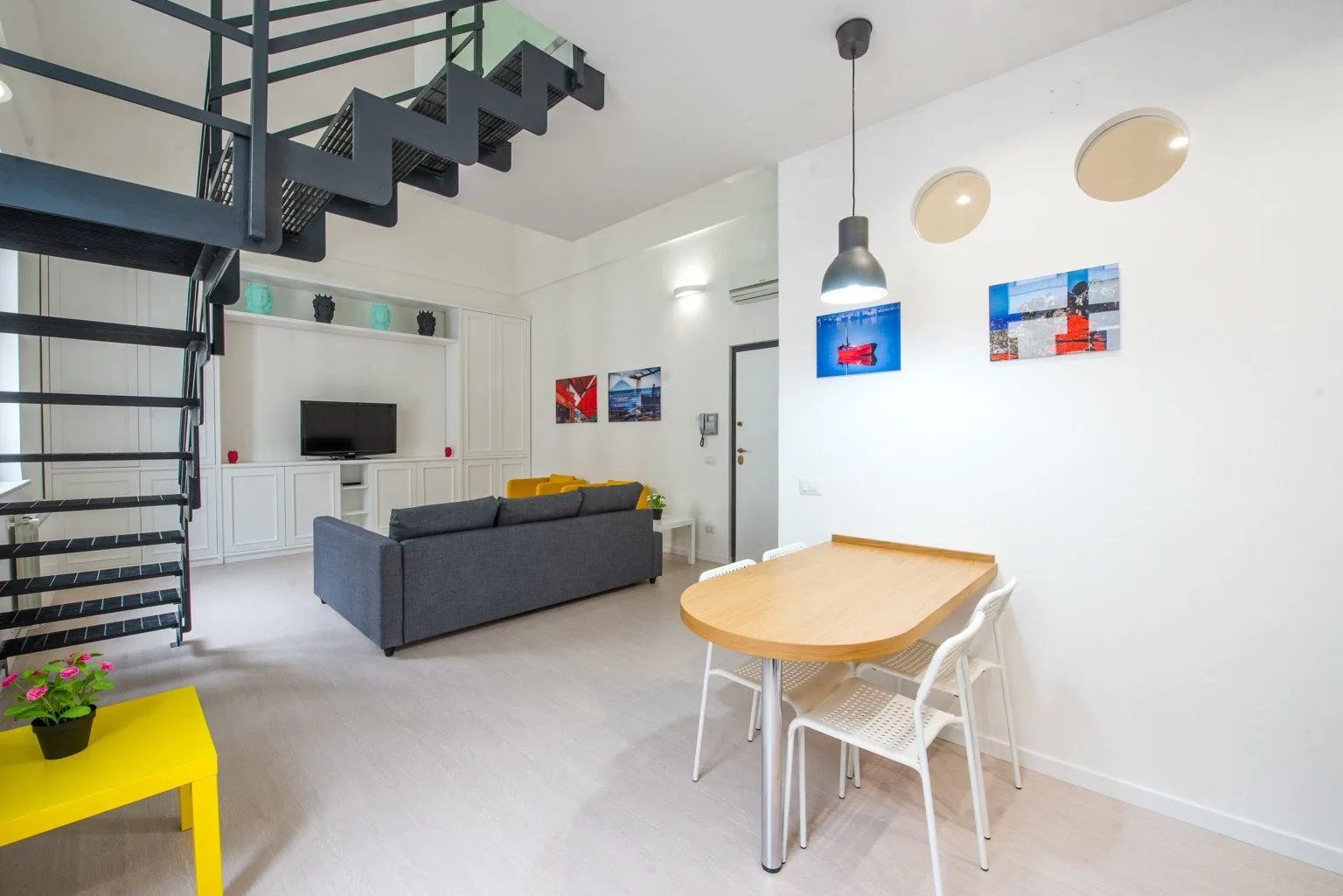 A bright living and dining area features a gray sofa, table, and open staircase.