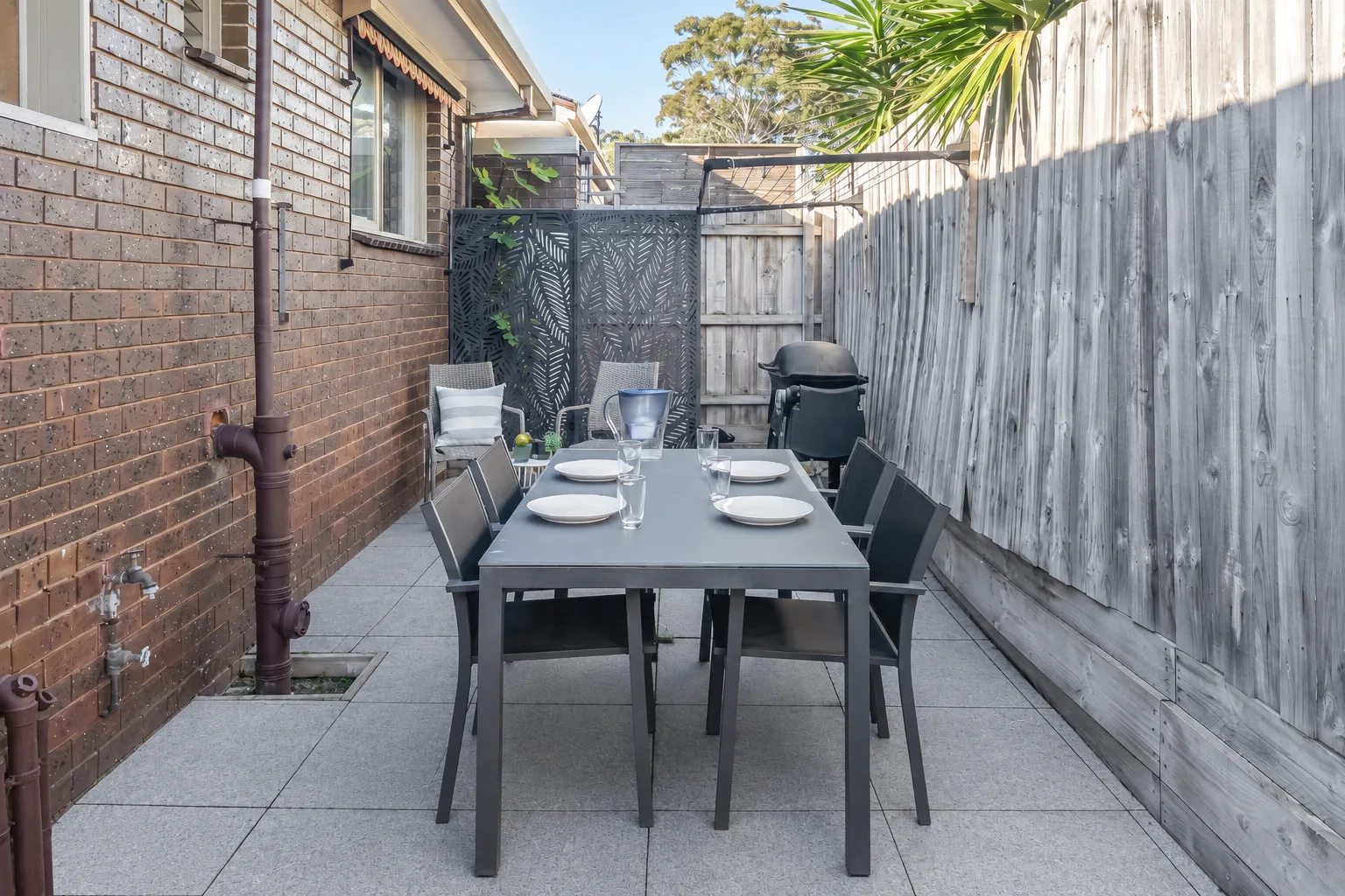 Una zona de comedor al aire libre tiene una mesa larga con sillas junto a una pared de ladrillo.