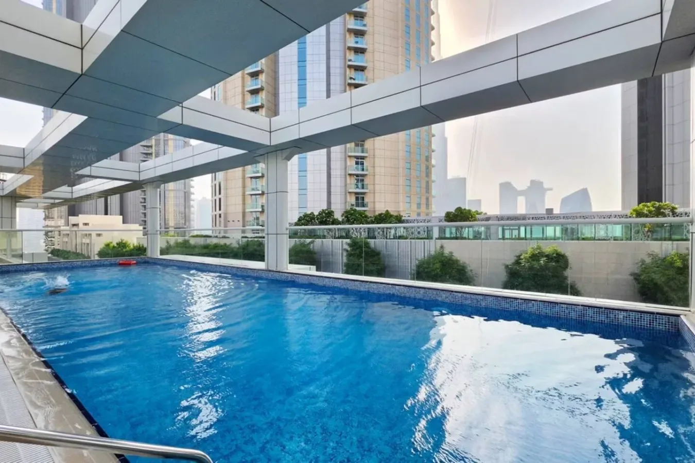 Una piscina al aire libre está bajo una cubierta geométrica, con edificios altos al fondo.