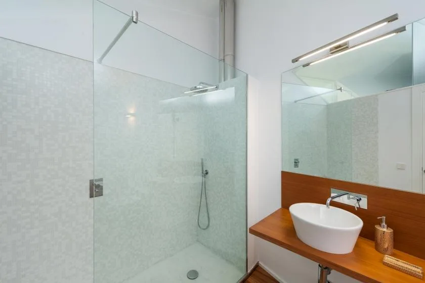 Un baño luminoso con una ducha de vidrio y un lavabo blanco sobre una encimera de madera.