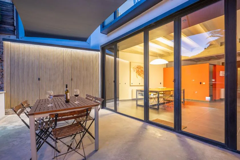 Una zona de comedor al aire libre da a un interior iluminado a través de grandes puertas de vidrio.