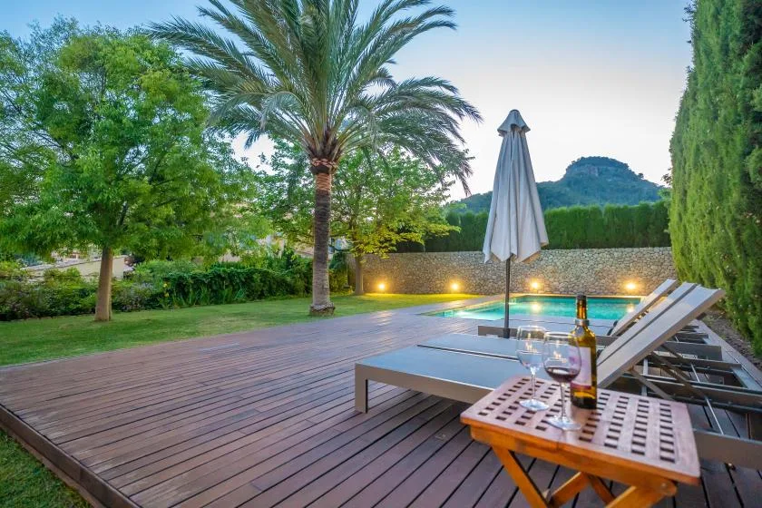 Una terraza junto a la piscina con asientos reclinables, una sombrilla cerrada y vegetación.