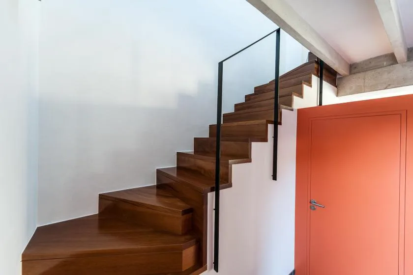 Una escalera de madera oscura con barandilla de vidrio sube junto a una puerta naranja.