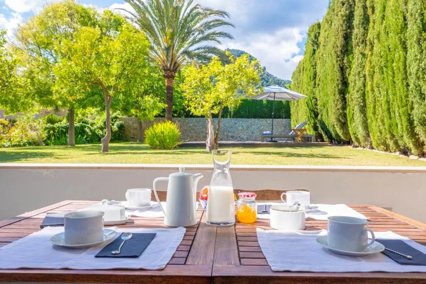 Una mesa al aire libre está puesta con tazas y una cafetera junto a un jardín.