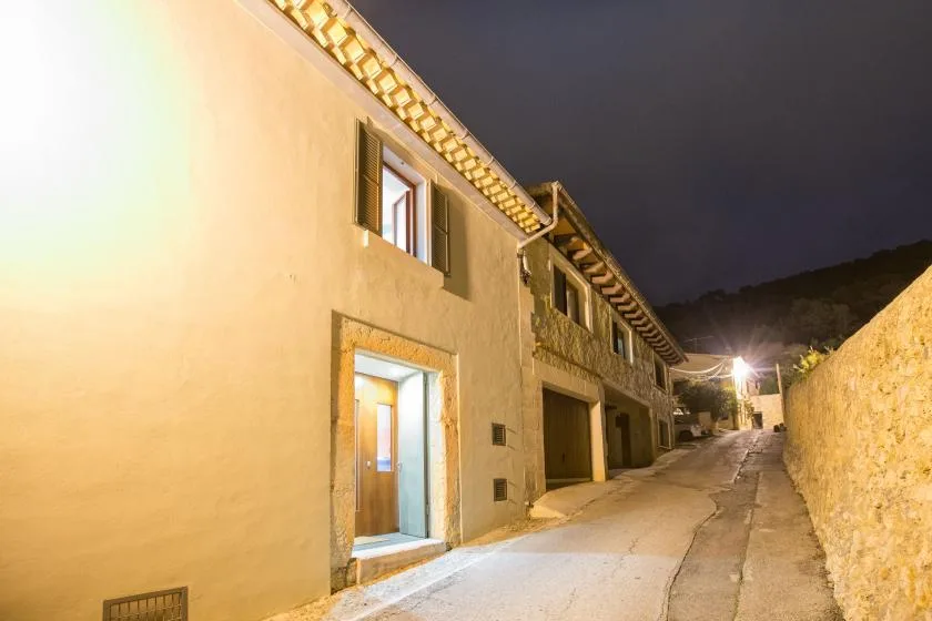 Un callejón pavimentado y estrecho pasa entre edificios iluminados y un muro de piedra por la noche.