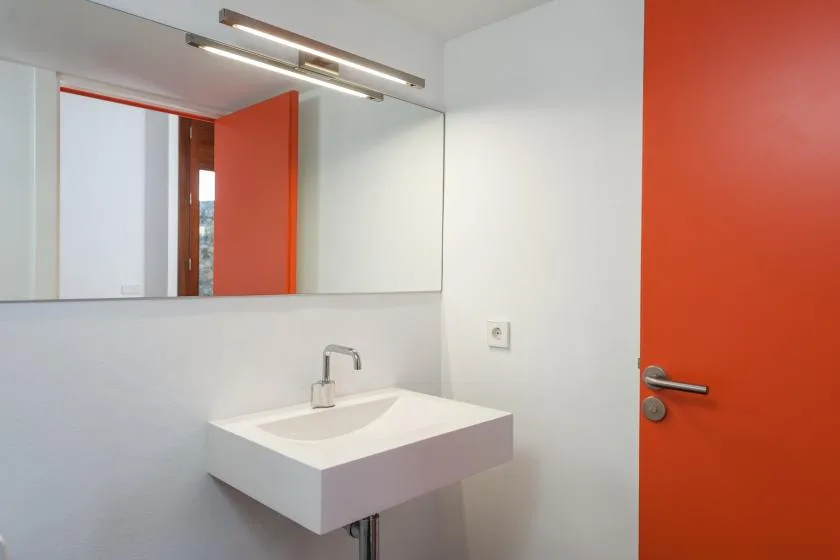 Un baño con lavabo suspendido, espejo grande y puerta roja.