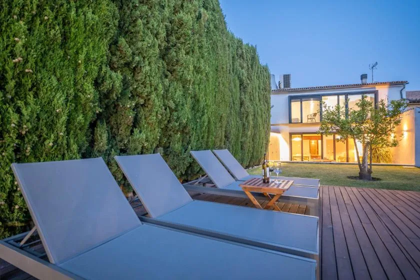 Zona de descanso junto a una terraza de madera, con una casa iluminada al fondo.