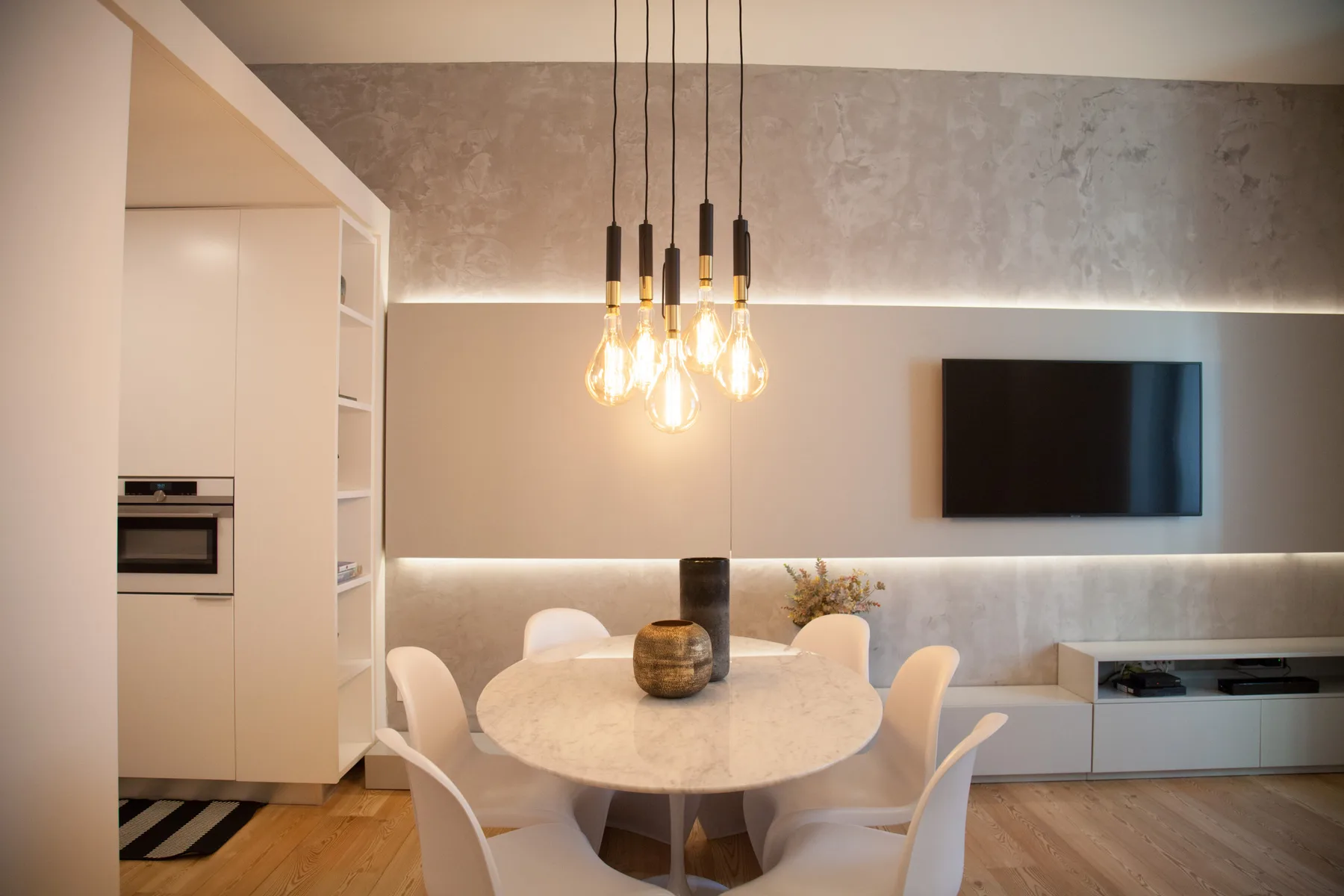 A modern dining area with a round table and pendant lights beside a wall-mounted television.