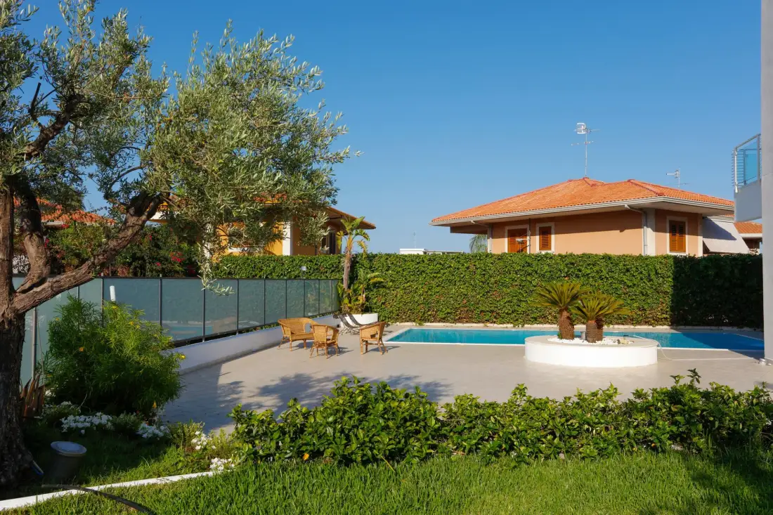 Una zona de piscina al aire libre con terraza pavimentada, vegetación y casas cercanas.