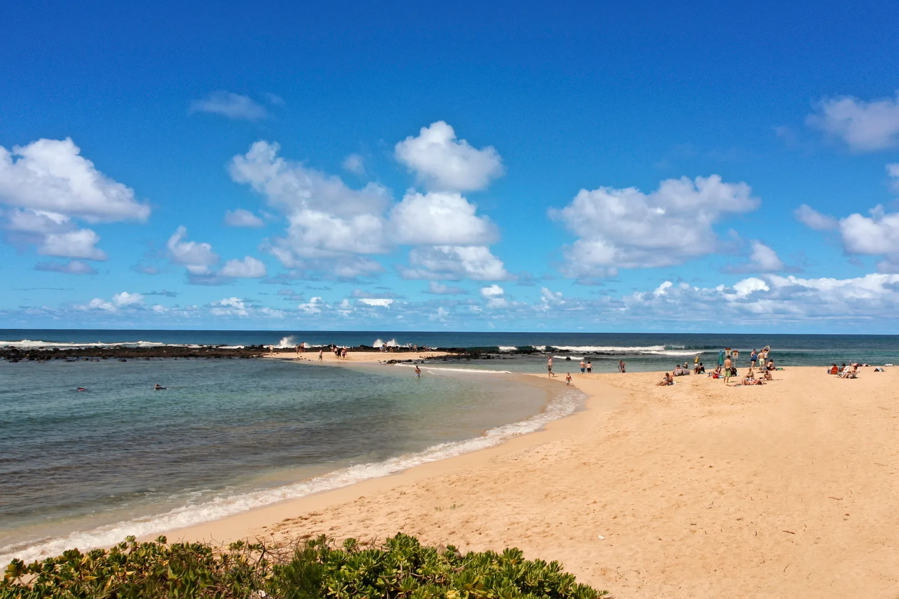 Una playa de arena se curva junto a aguas poco profundas bajo un cielo azul con nubes.