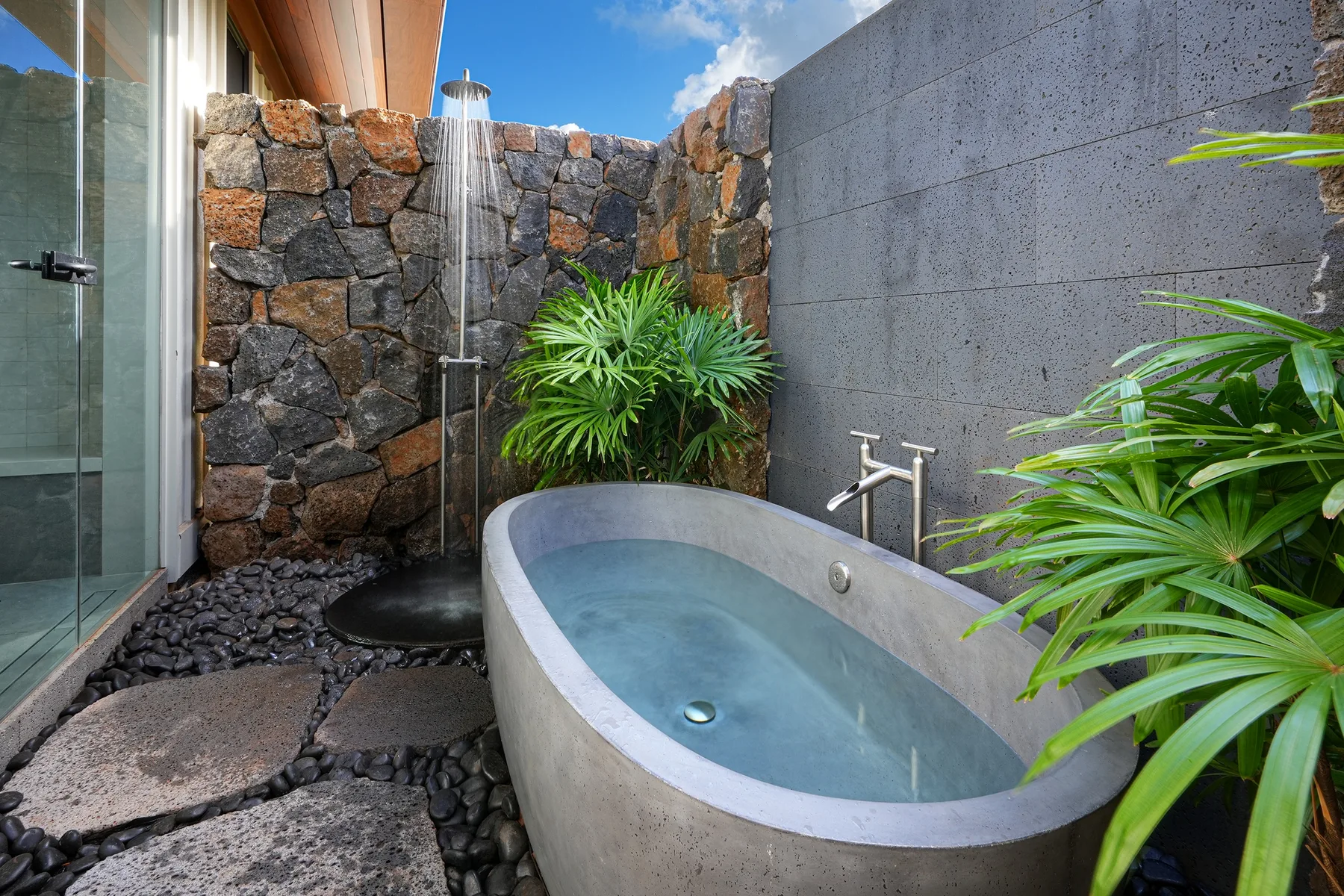 Una zona de baño al aire libre con bañera, ducha y plantas tropicales.