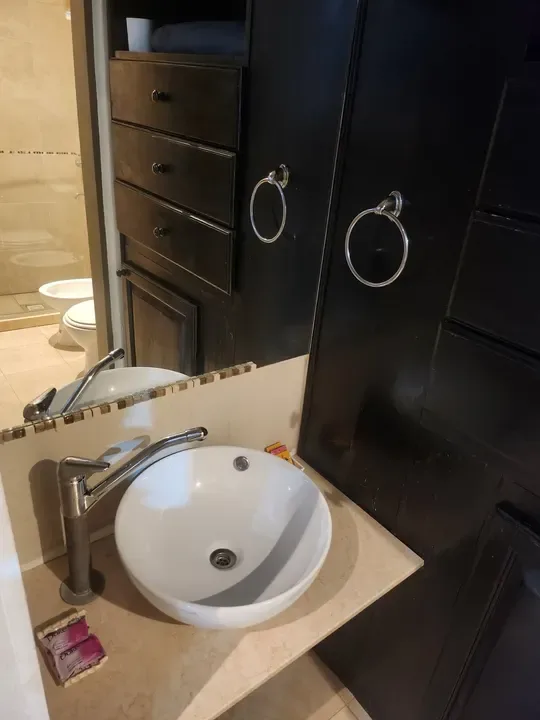 Lavabo blanco sobre una encimera clara, espejo, mueble oscuro y ducha al fondo.