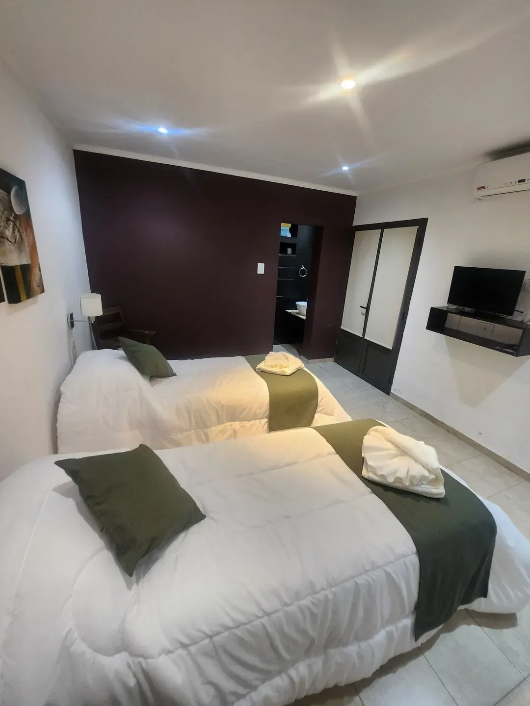Dos camas individuales con detalles verdes, un televisor y la puerta del baño al fondo.