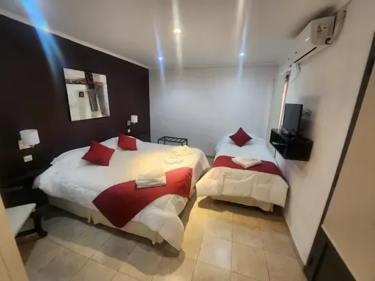 Tres camas individuales, dos de ellas juntas, con cojines rojos, un televisor y aire acondicionado.