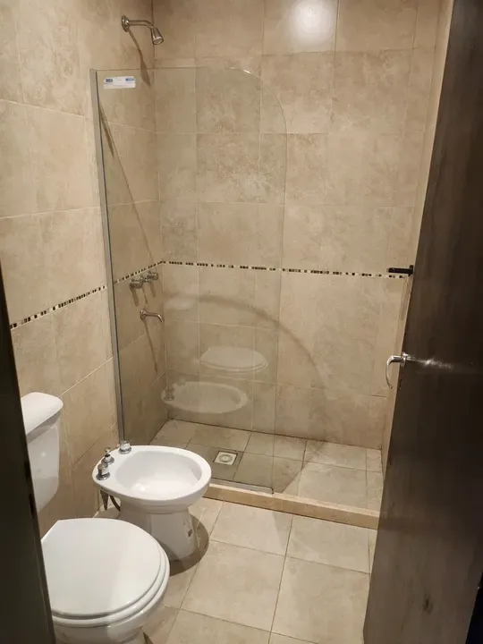 Baño con mampara de ducha de vidrio, inodoro y bidé, en tonos claros.