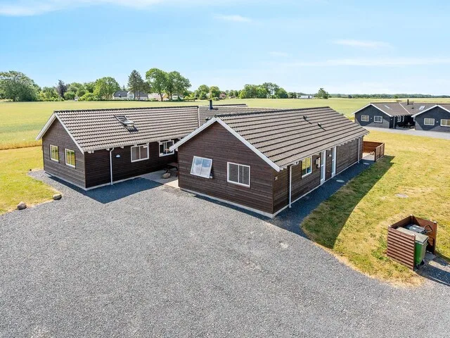 A dark wooden vacation house stands beside a gravel driveway and open grassy fields.